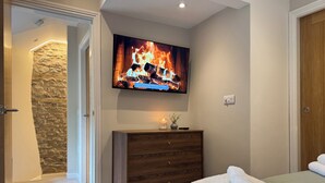A bedroom with a bed, a dresser, a TV displaying a fire, and a stone wall. - Railway Cottage (Oswestry)