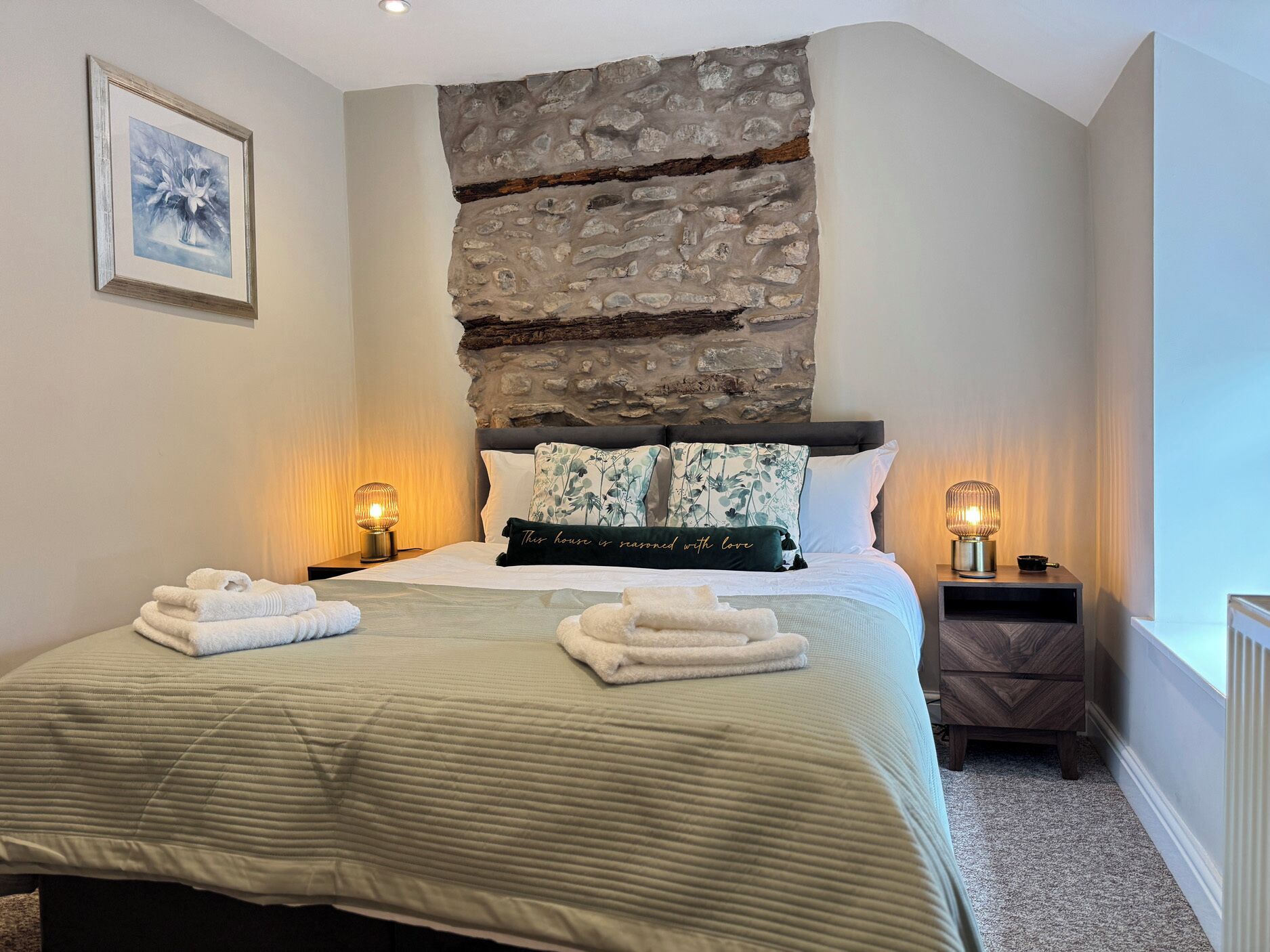 A bedroom with a stone wall, a bed with multiple pillows, two bedside lamps, and a framed picture on the wall.
