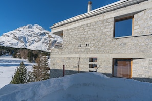 Ein Steinhaus mit großem Fenster, Schornstein und verschneiter Landschaft, im Hintergrund sind Berge zu sehen.