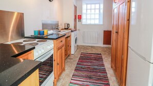 A compact kitchen with a stove, sink, and refrigerator, wooden cabinets, a clock, and a window. - The Old Coach House (Lochmaben)