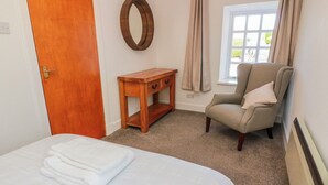 A hotel room with a bed, a wooden nightstand, a chair, a window with curtains, and a wall-mounted mirror. - The Old Coach House (Lochmaben)