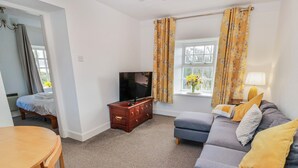 A living room with a sofa, a TV, a wooden cabinet, a dining table, and a window with curtains. - The Old Coach House (Lochmaben)