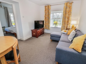A living room with a sofa, a TV, a wooden cabinet, a dining table, and a window with curtains. - The Old Coach House (Lochmaben)