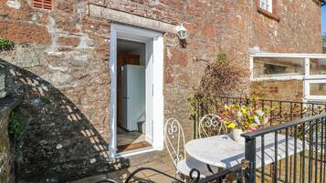 A stone building with a white door, a small balcony with a table and chair, and a metal railing.