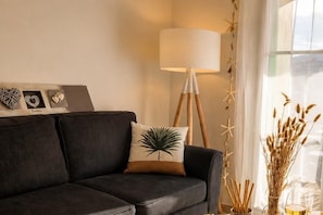 Un canapé en cuir foncé agrémenté d’un coussin à motifs de feuilles de palmier, une lampe sur pied et des objets décoratifs disposés sur une étagère.