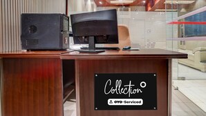 Um balcão de madeira na recepção, com um computador, uma placa indicando 'Collection' e 'OYO-Serviced', e um interior moderno com ventoinha no teto e divisórias de vidro.