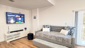 Un salon moderne avec un canapé gris, un téléviseur à écran plat fixé au mur, un meuble de rangement blanc et une fenêtre équipée de stores.