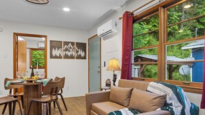 A cozy room with a dining table, chairs, a sofa, a lamp, and a large window with a view of trees. - Bryson City Cabin · Sleeps 5 · Pets OK · Fire Pit (Whittier)