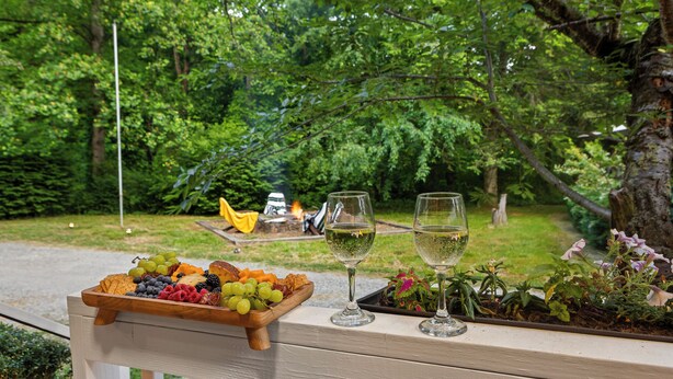 A wooden bench with a fruit platter and two glasses of white wine, overlooking a lush garden with a fire pit. - Bryson City Cabin · Sleeps 5 · Pets OK · Fire Pit (Whittier)