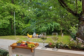 Een houten bank met een fruitschaal en twee glazen witte wijn, met uitzicht op een weelderige tuin met een vuurkorf.