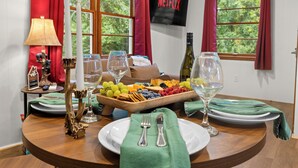 A dining table with a fruit platter, candles, wine glasses, and cutlery, set against a window with red curtains and a TV displaying 'NETFLIX'. - Bryson City Cabin · Sleeps 5 · Pets OK · Fire Pit (Whittier)