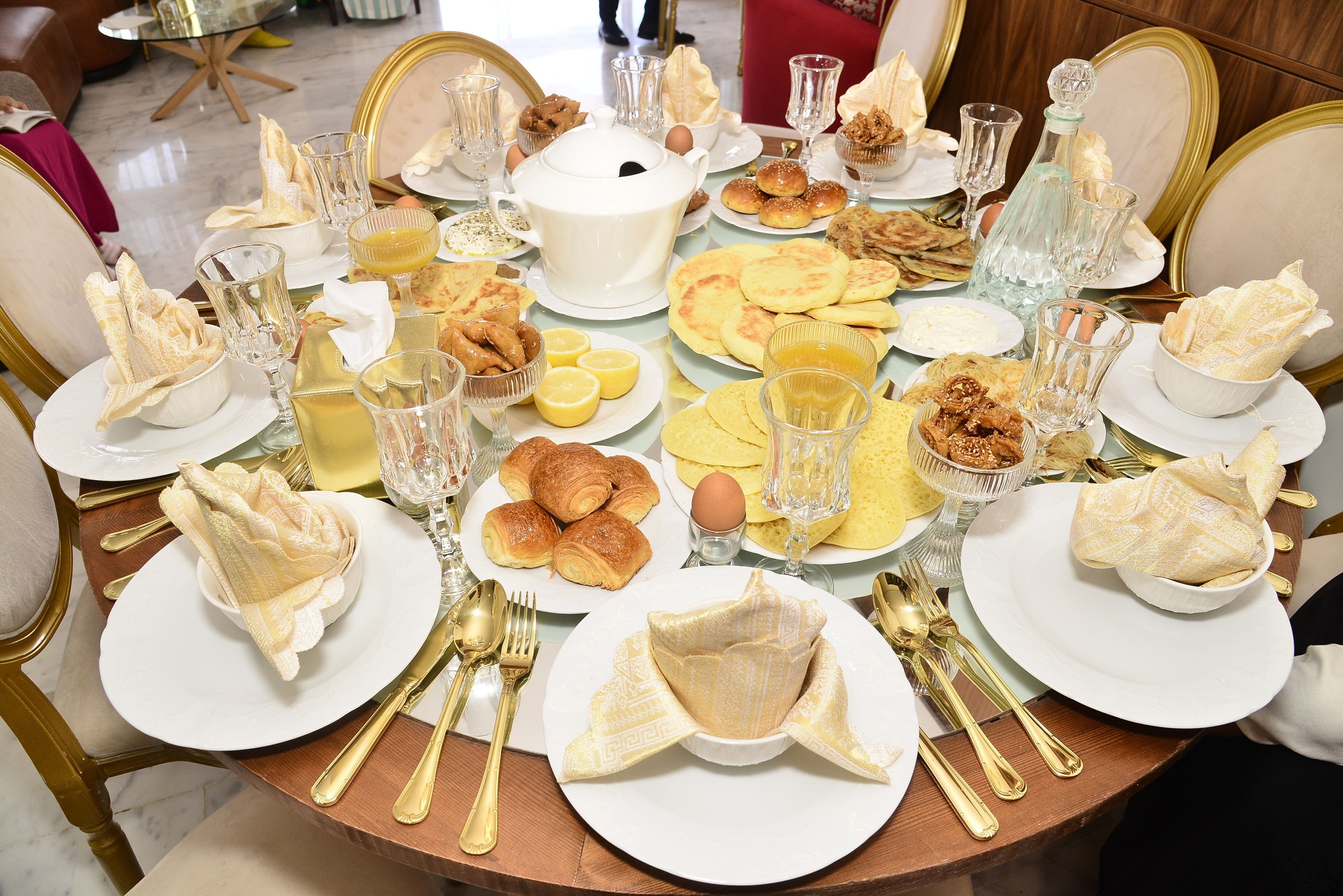Una mesa de comedor redonda con platos blancos, cubiertos dorados y diversos alimentos.