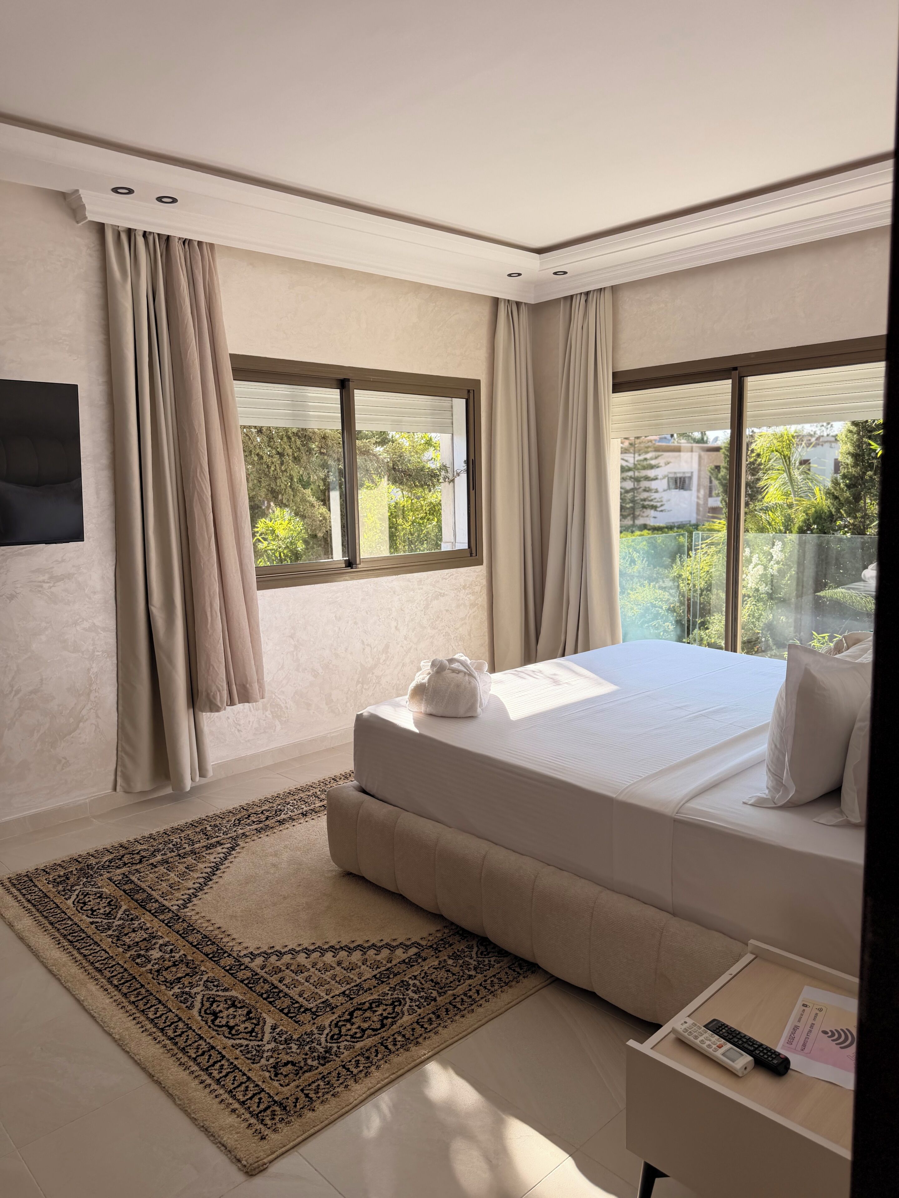 A modern bedroom with a large bed, a flat-screen TV, and a view of greenery outside.