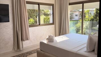 A modern bedroom with a large bed, a flat-screen TV, and a view of greenery outside.