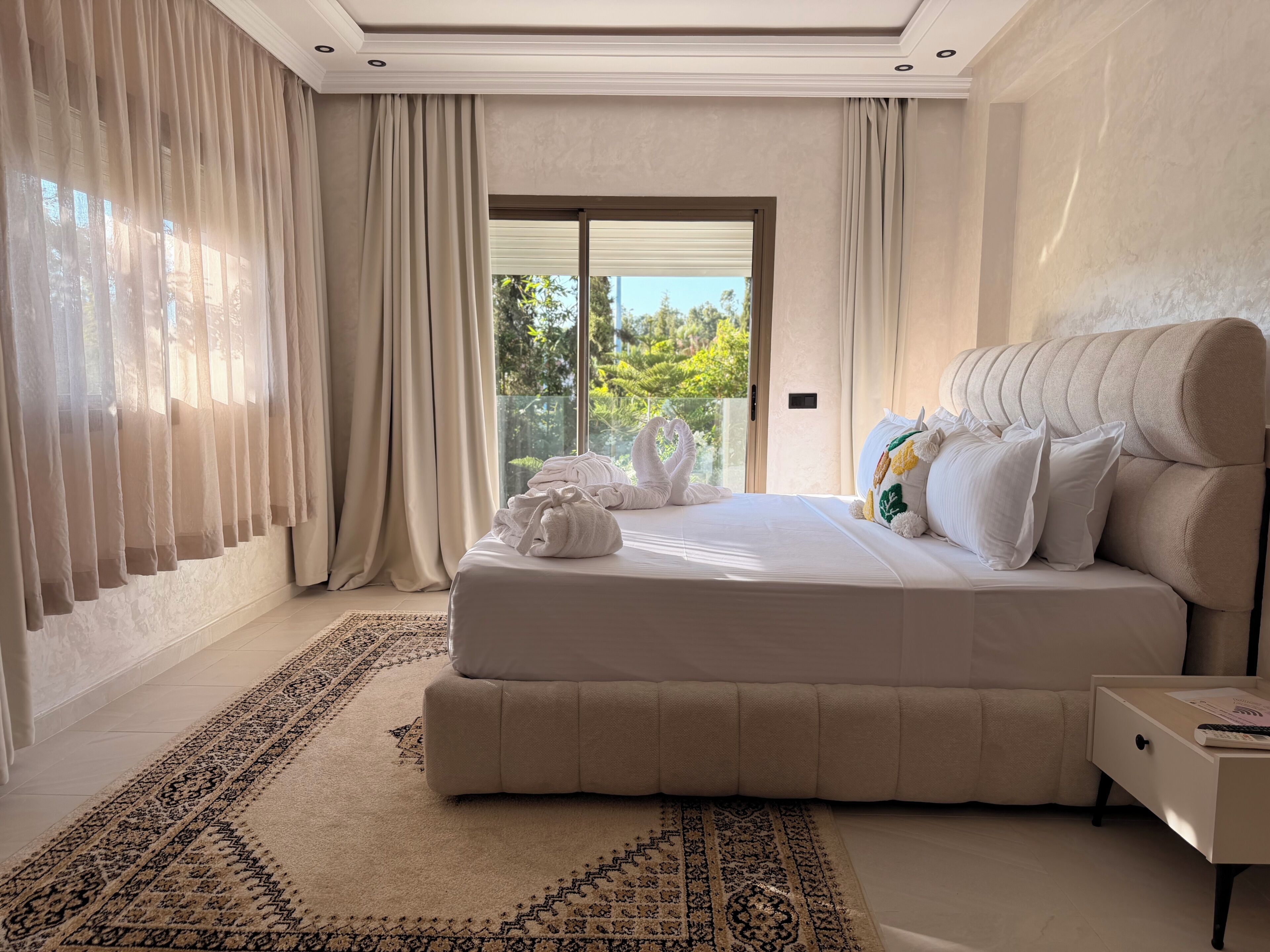 Un dormitorio con una cama grande, una mesita de noche, un tapete estampado y vistas al verdor exterior.