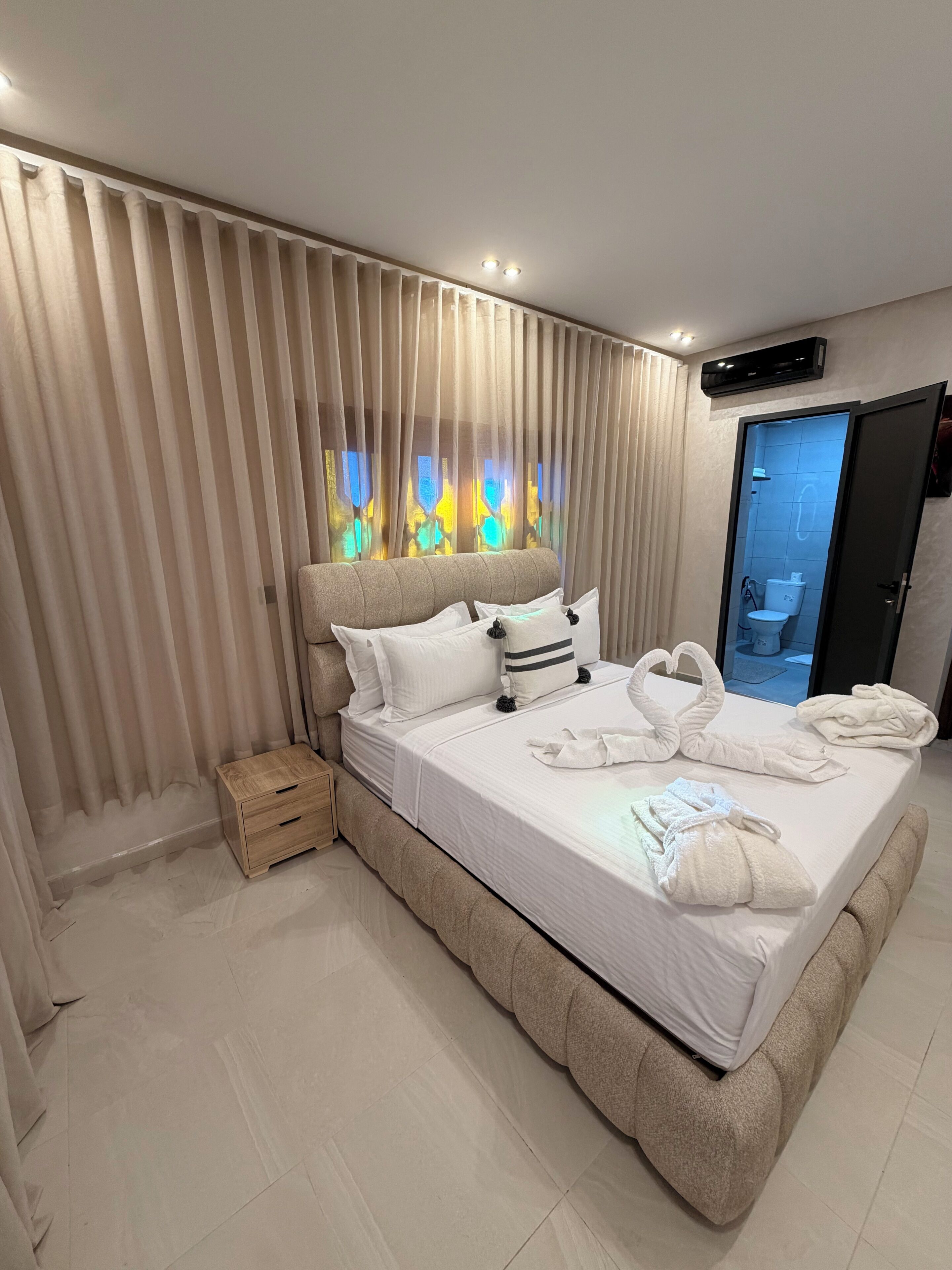A modern hotel room with a large bed, a wooden nightstand, and a bathroom visible through a glass door.