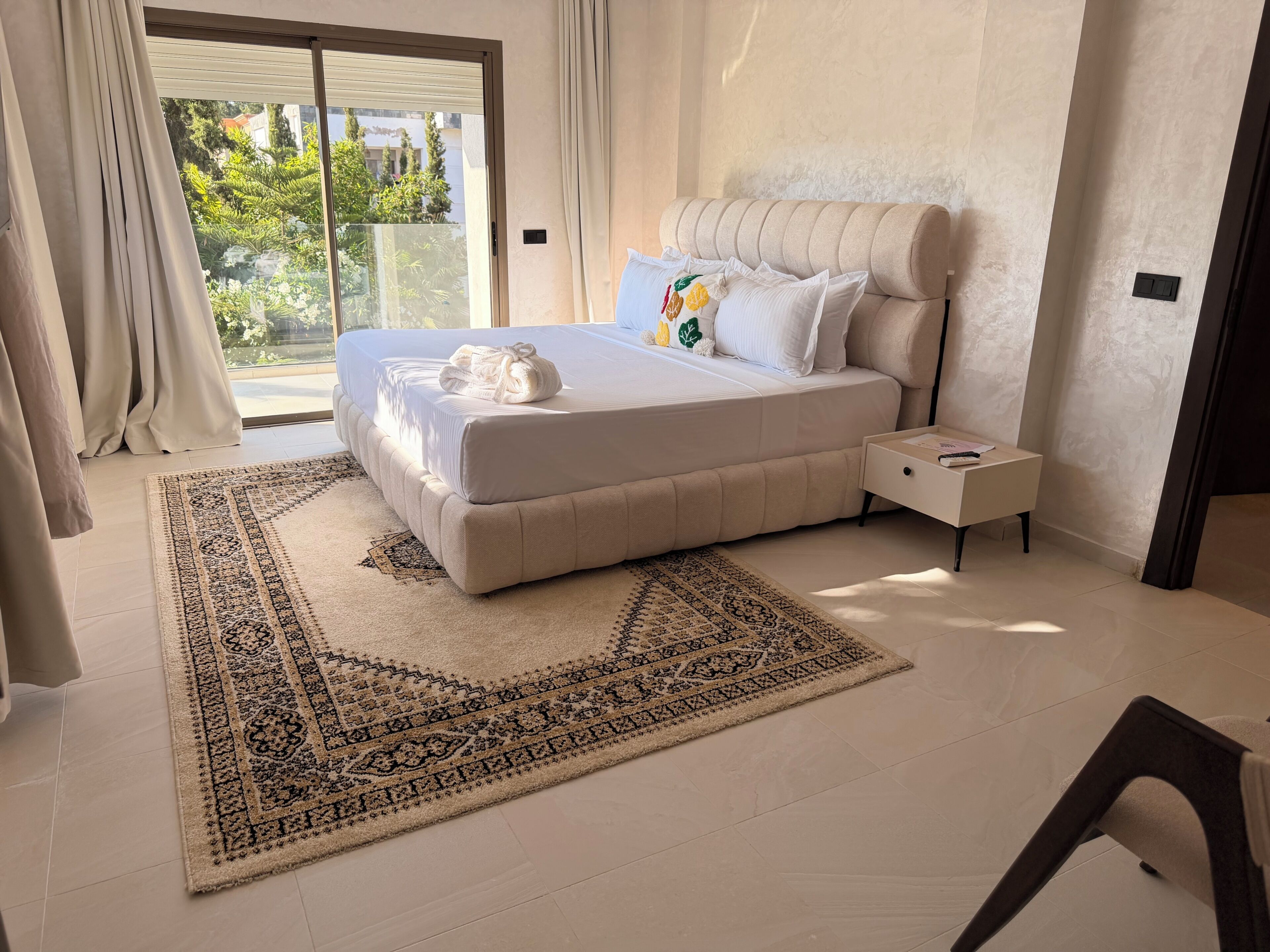 A modern bedroom with a large bed, a nightstand, a patterned rug, and a view of greenery outside.