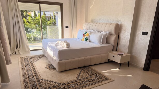 A modern bedroom with a large bed, a nightstand, a patterned rug, and a view of greenery outside. - Riad Elizabeth (Nouaceur)