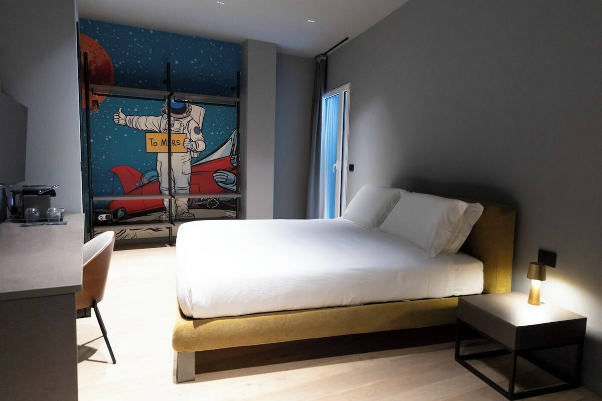 Quarto de hotel moderno com uma cama, uma mesa de escritório e um mural de astronautas.