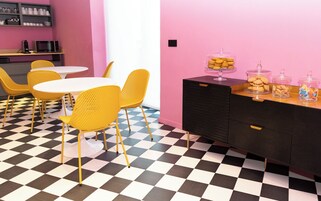 Una habitación con suelo a cuadros, sillas amarillas, una mesa blanca, un mueble negro con cajones y una pared rosa.