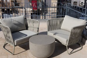 A patio with two grey wicker chairs and a round table, surrounded by a metal fence.