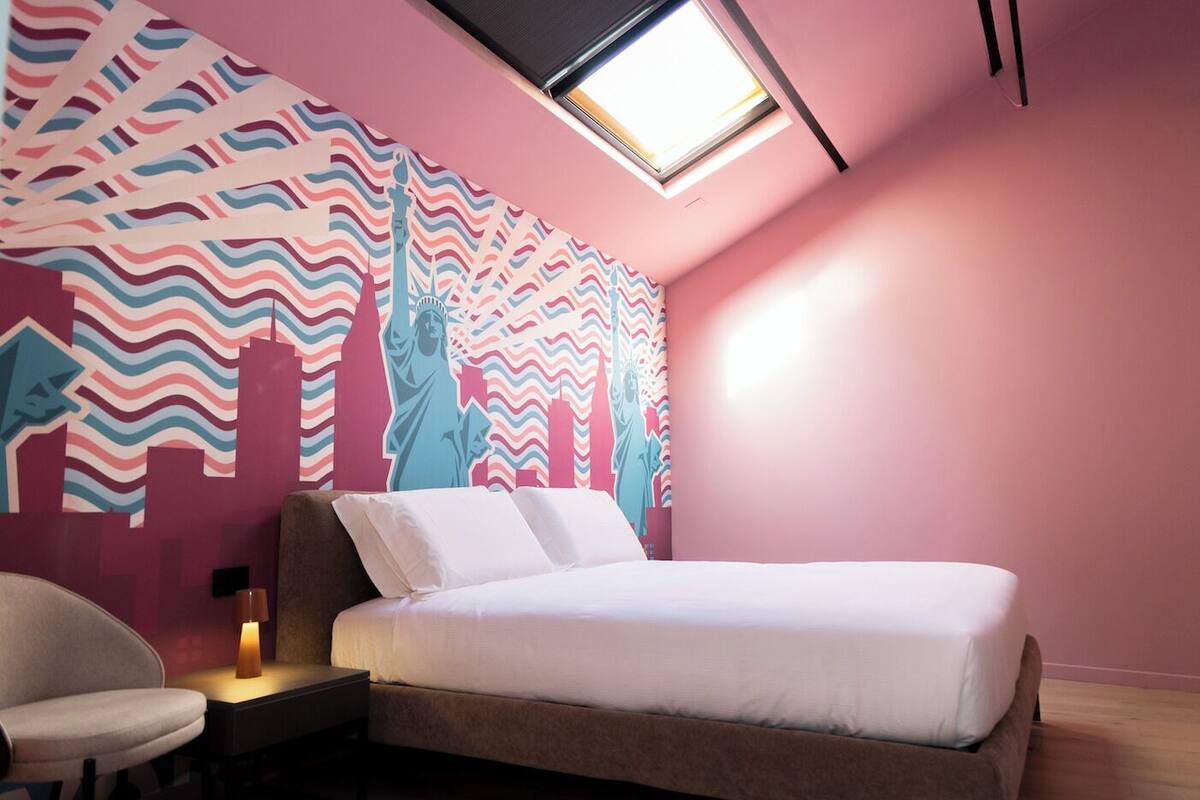 Quarto com cama, cadeira, mesa de cabeceira e parede com mural da paisagem urbana.
