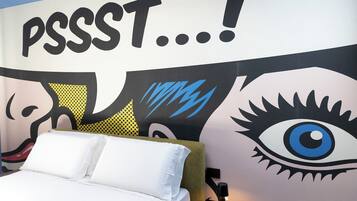 Une chambre à coucher avec un lit, des oreillers, une table de chevet et un mur orné d’un grand motif représentant un œil et d’œuvres d’art abstraites.