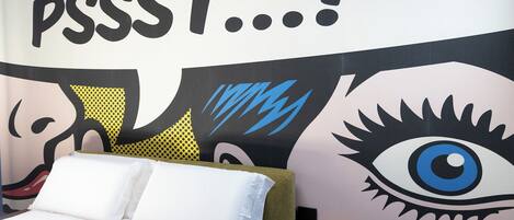Une chambre à coucher avec un lit, des oreillers, une table de chevet et un mur orné d’un grand motif représentant un œil et d’œuvres d’art abstraites.