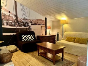 A living room with a sofa, a coffee table, a lamp, a patterned cushion, and a wall with a nautical-themed mural. - Charming Charentaise house, ideal for peaceful vacations. (Saint-Palais-sur-Mer)