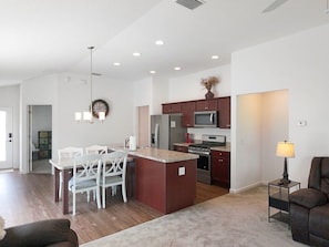 A modern kitchen with a central island, dining area with white chairs, and a living room with a dark sofa. - Meadow View - In The Villages (Wildwood)