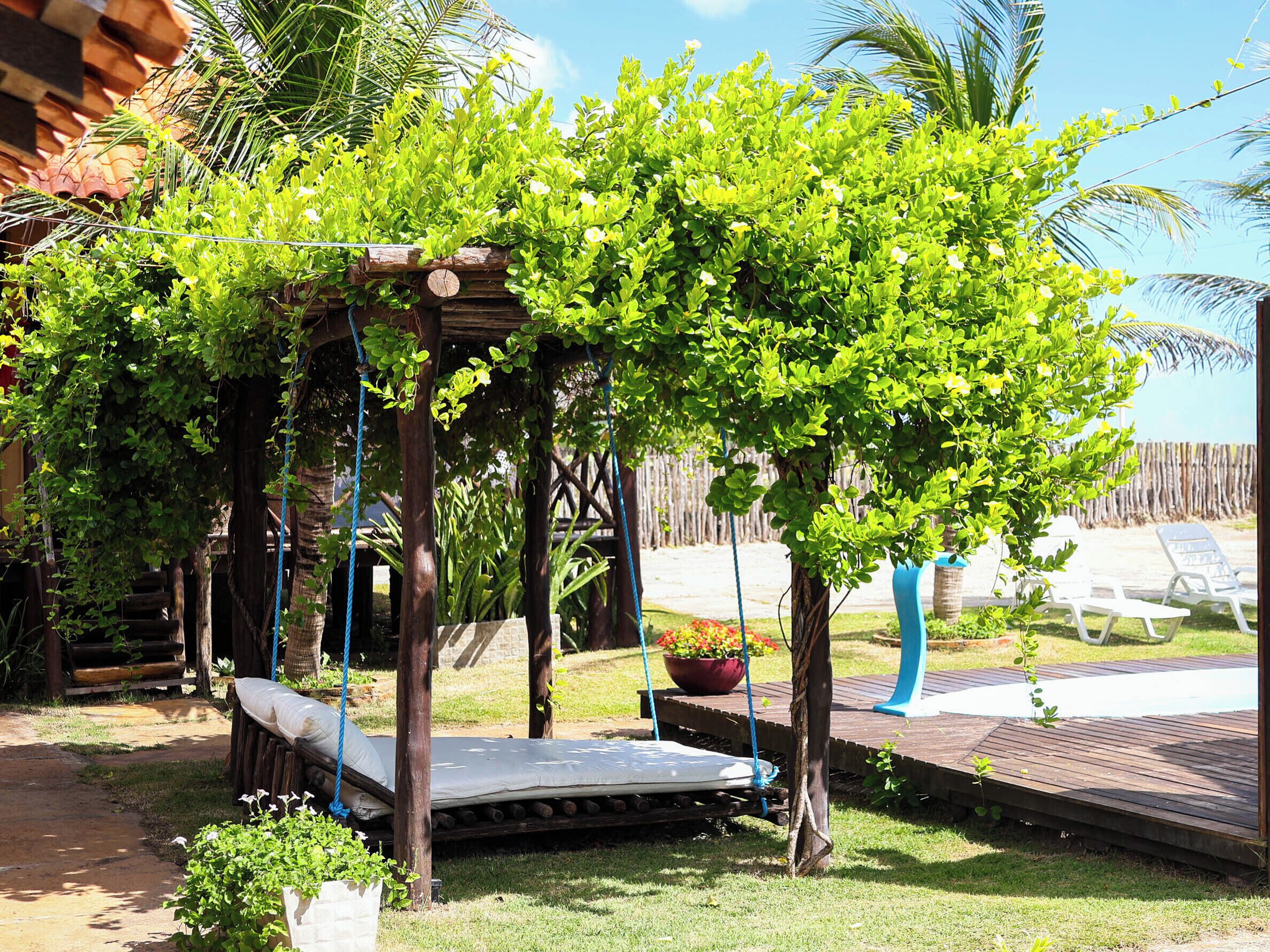 Una pérgola de madera con una hamaca, un pequeño trampolín y un jardín con plantas en macetas y una palmera.