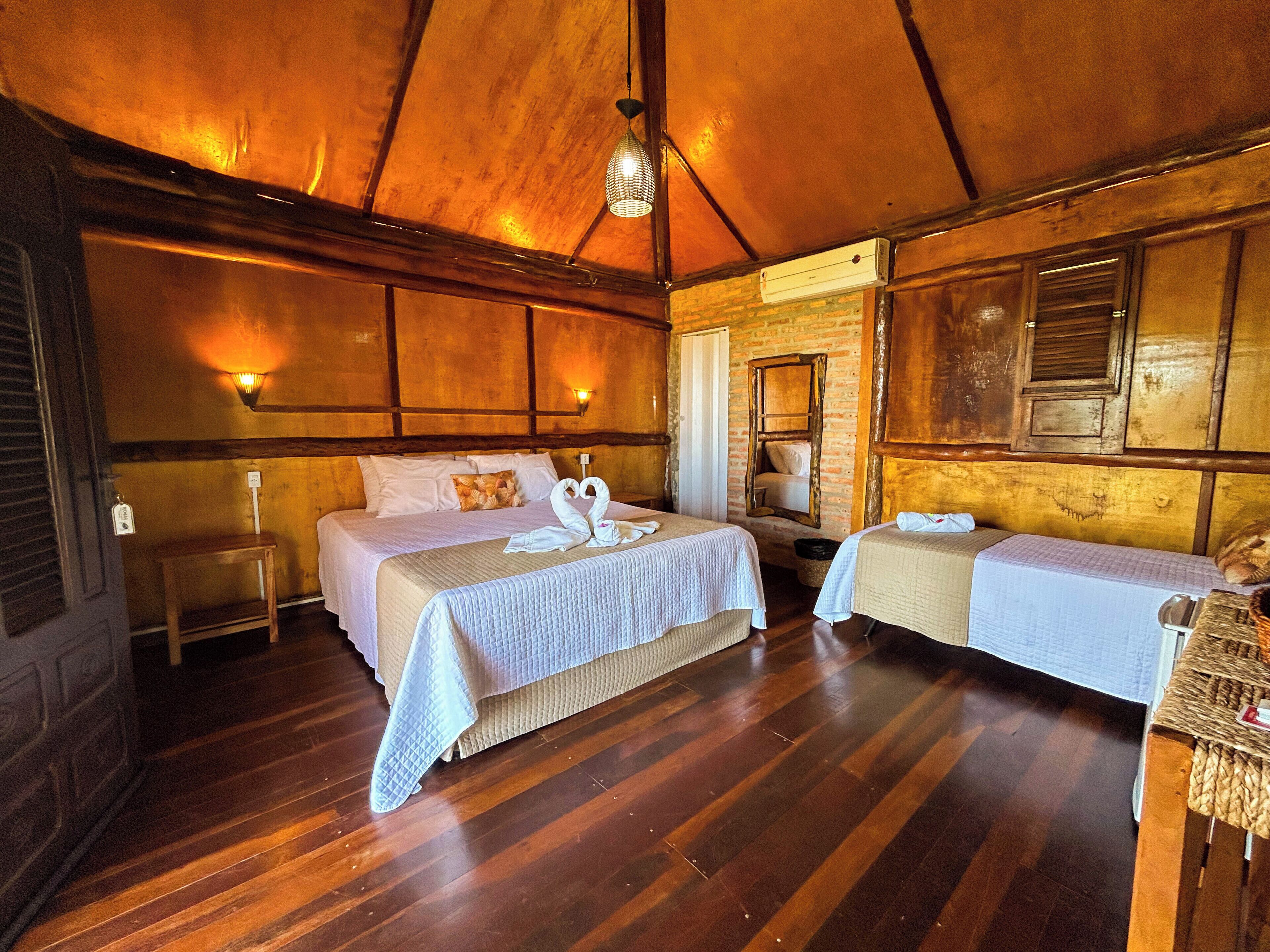Een kamer met een houten plafond, twee bedden met witte lakens, een hanglamp en een bakstenen muur.