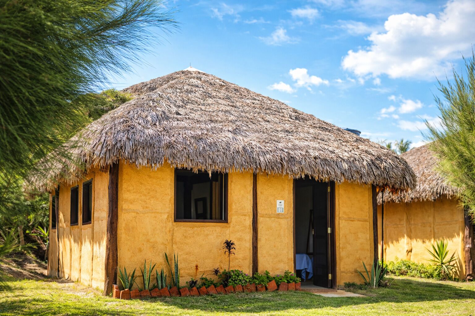 Una cabaña de techo de paja con una ventana y una puerta visibles, rodeada de un jardín bien cuidado con diversas plantas.