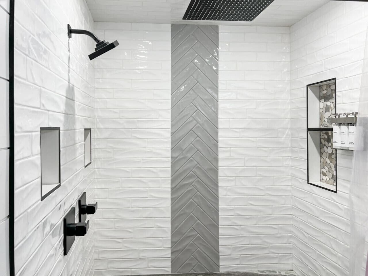 A modern bathroom with white tiled walls, a herringbone-patterned column, a showerhead, and a framed picture with toiletries.