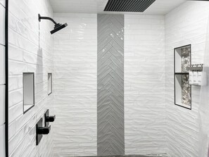 A modern bathroom with white tiled walls, a herringbone-patterned column, a showerhead, and a framed picture with toiletries.