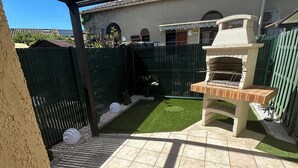 Eine Terrasse mit Grill, grünem Zaun und einem Haus im Hintergrund.