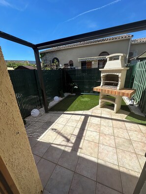 Eine Terrasse mit Grill, grünem Zaun und einem Haus im Hintergrund.