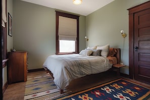 Ein Schlafzimmer mit einem Bett, einer Holzkommode, einem Fenster mit Jalousien, einer Wandleuchte und einer Tür.
