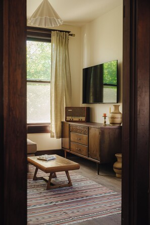 Ein Zimmer mit einem Holzkabinent, einem an der Wand montierten Fernseher, einem Fenster mit Vorhängen und einem gemusterten Teppich.