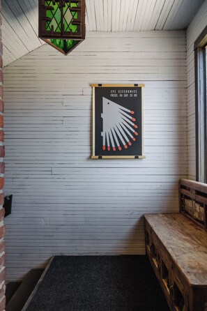 Ein Zimmer mit weißen Holzwänden, einer schwarzen Matte am Eingang, einer hölzernen Bank und einem gerahmten Bild mit geometrischem Muster.