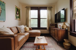 A living room with a brown leather sofa, a wooden coffee table, a flat-screen TV, and a window with curtains. - The Apartment at Lucky Duck - 2 bedrooms in a historic restored bank (Carlton)