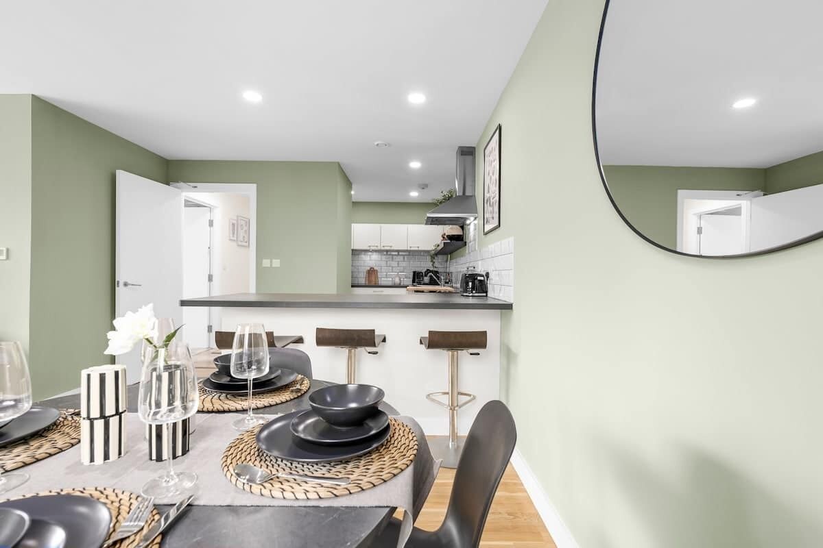 A dining area with a table set for four, a kitchen in the background, and a mirror on the wall.