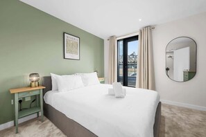 Ein Hotelzimmer mit Bett, Nachttisch, Wandspiegel und einem fensterbelaubten Fenster.