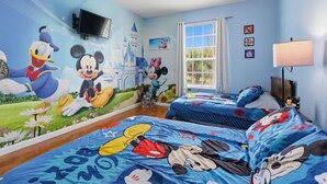 Et børneværelse med vægmalerier af Mickey Mouse og Donald Duck, to senge med blåt sengetøj, et fjernsyn og et vindue med gardiner.