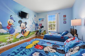 Et børneværelse med vægmalerier af Mickey Mouse og Donald Duck, to senge med blåt sengetøj, et fjernsyn og et vindue med gardiner.