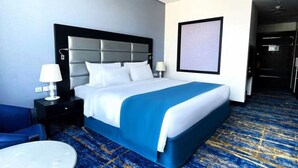 A hotel room with a large bed, two bedside tables with lamps, a blue carpet, and a door leading to another room. - Classic Room | Sig1 Barsha | Rooftop Pool (Dubai)
