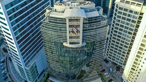 A high-rise building with a distinctive curved facade, a prominent logo on the top, and a parking area in front. - Classic Room | Sig1 Barsha | Rooftop Pool (Dubai)