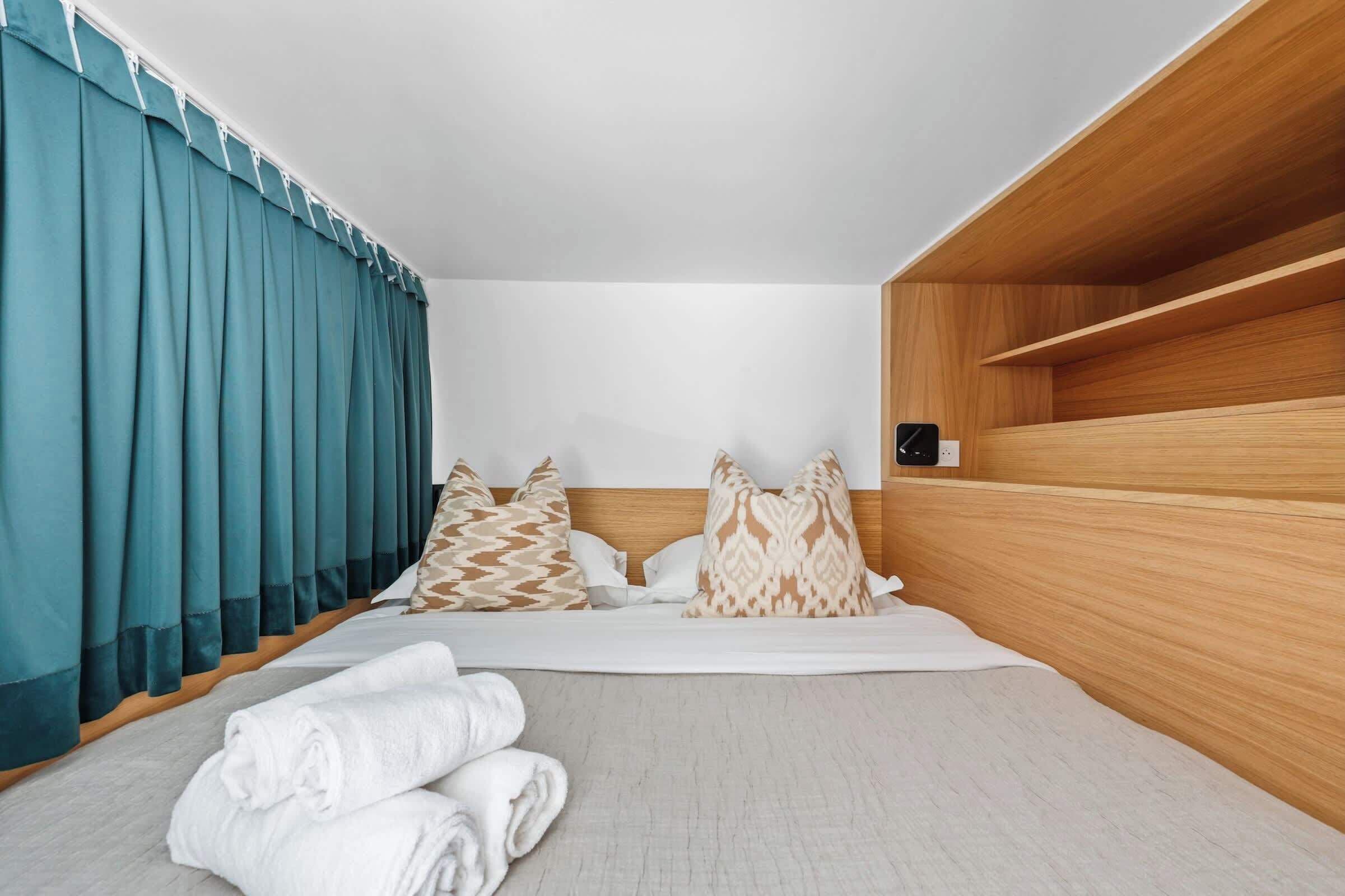 A hotel room with a bed, pillows, a wooden shelf, and a window with curtains.