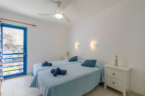 Een slaapkamer met twee bedden, een plafondventilator, bedlampjes, een nachtkastje en uitzicht door een deur met blauwe lijst.