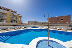 Een zwembad met ligbedden, een jacuzzi en een gebouw met balkons op de achtergrond.
